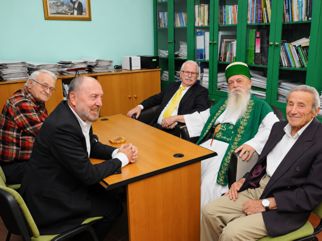 Robert Ince and Bektashi hosts sit around a desk in an office with green bookshelves.